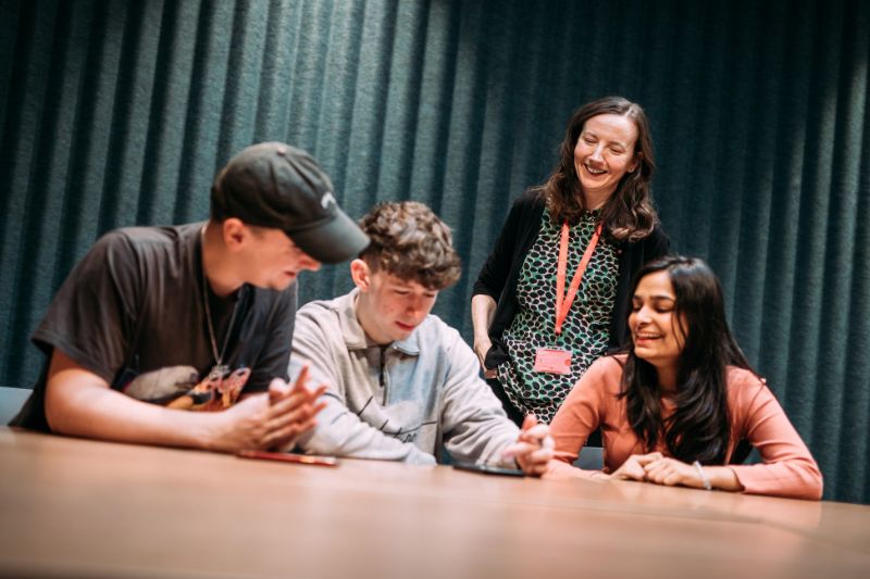 Three students interacting together at a desk with a staff member looking on and smiling